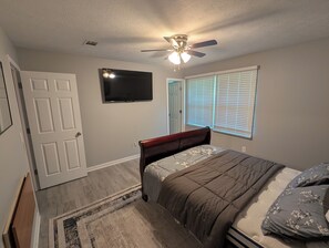 A bedroom with a bed, a ceiling fan, a TV, a door, and a window with blinds.