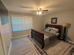 Un dormitorio con cama, ventilador de techo, ventana con persianas, alfombra y un cuadro en la pared.