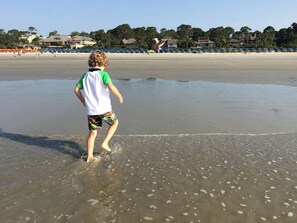 A beach with a child walking in shallow water, a row of blue umbrellas, and buildings in the background. - Available 6/26-7/3/26 only! Sea Pines Villa; 2BR/2BA; Sleeps 6; HHI (Hilton Head Island)