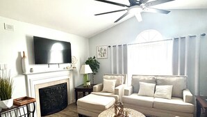 Un salon doté d’une cheminée, d’un téléviseur, d’un ventilateur de plafond et d’une grande fenêtre avec des rideaux.