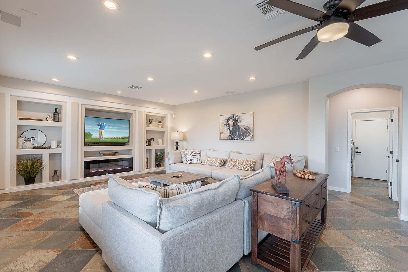 A living room with a sectional sofa, a flat-screen TV, built-in shelving, a ceiling fan, and a door leading to another room.
