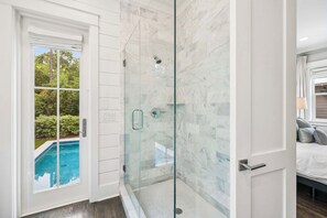 A modern bathroom with a glass shower enclosure, a visible pool outside, and a bed with a view.