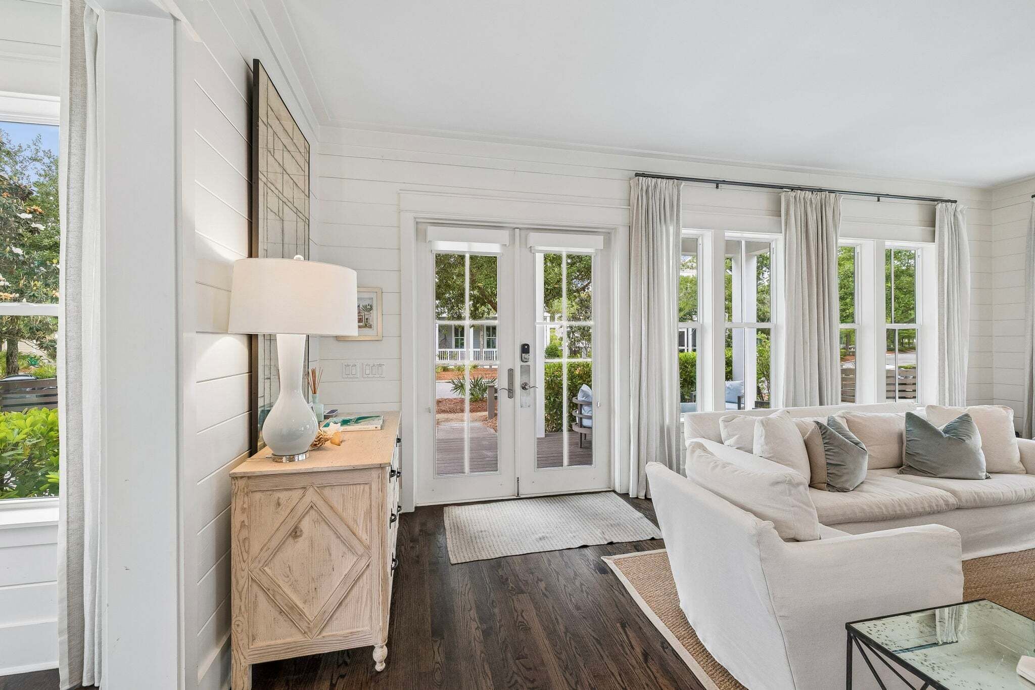 A room with a white sofa, a small wooden side table, a lamp, and a view through a glass door to an outdoor area.