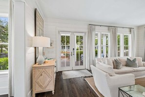 A room with a white sofa, a small wooden side table, a lamp, and a view through a glass door to an outdoor area.