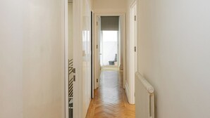 En smal hall med vita väggar och trägolv, som leder in till ett rum med ett stort fönster och ett element.