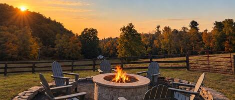 A circular stone fire pit with four Adirondack chairs around it, set in a grassy area with a wooden fence in the background.