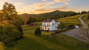 A large white house with a porch, surrounded by a fenced property, set in a rural landscape with hills in the background. - Heritage House - 13m to Shenandoah NP, Pool, Hot Tub, Mountain Views, EV (Page County)
