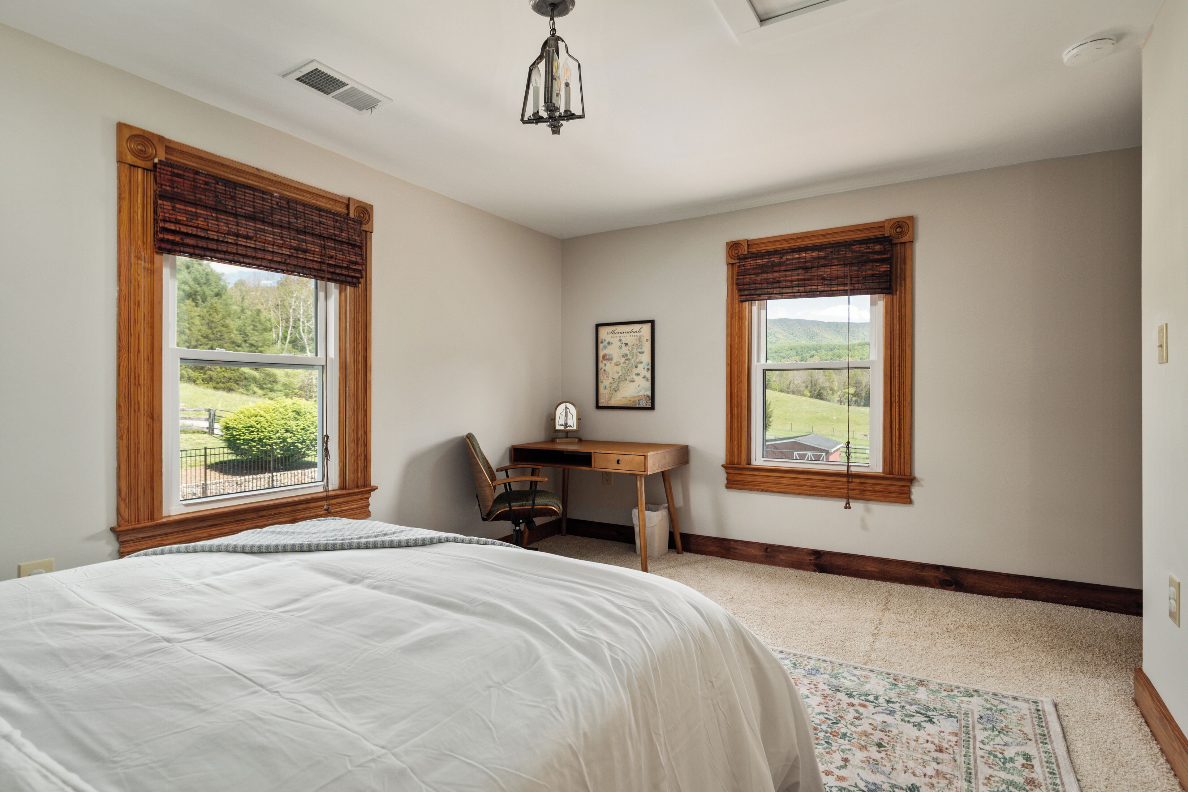 Ein Schlafzimmer mit einem Bett, einem Schreibtisch und zwei Fenstern mit Blick auf eine Landschaft.