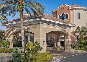 Ein Hotel im mediterranen Baustil, mit einem zentralen Eingangsbereich mit Bögen, einem Ziegeldach und einem angelegten Garten mit Palmen.