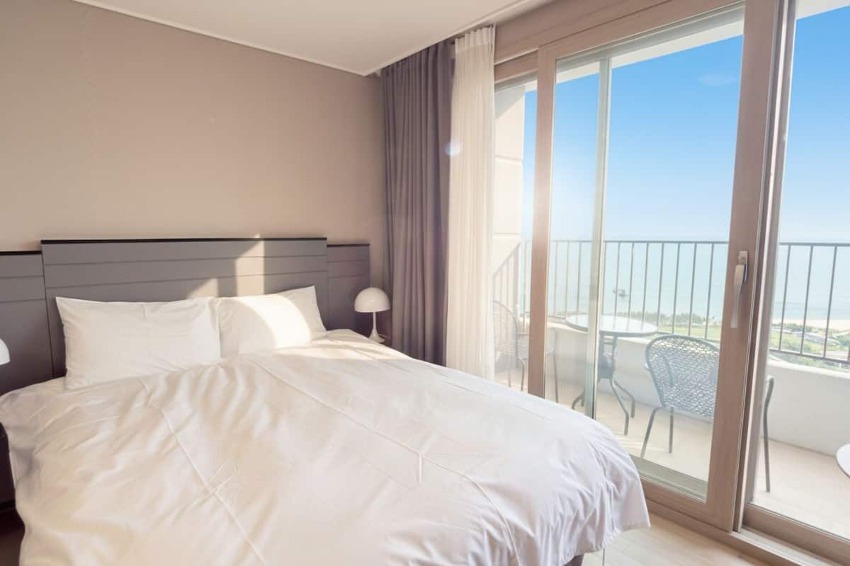 Habitación de hotel con cama, mesita de noche, lámpara, balcón con una silla y vista al mar.