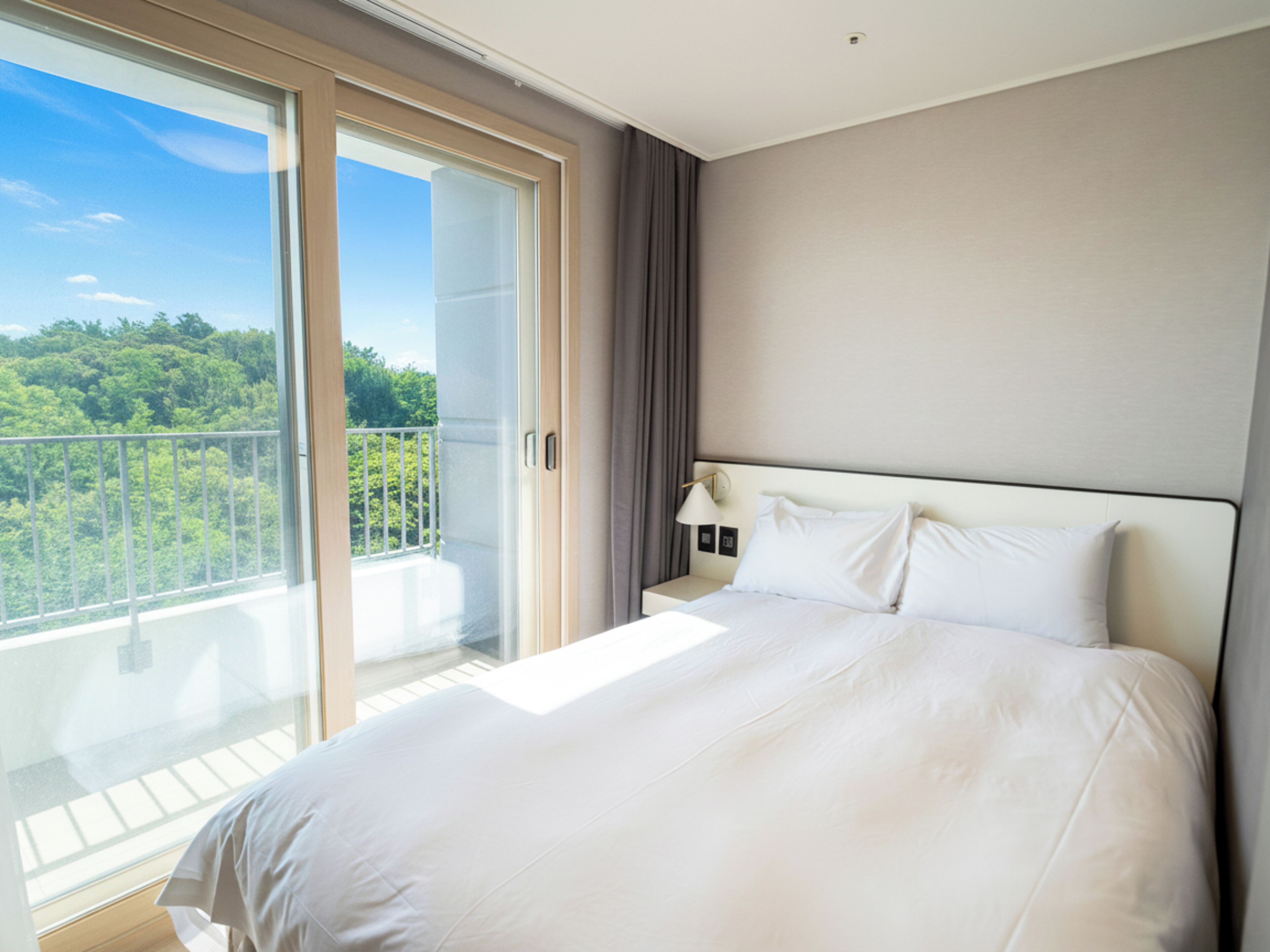 A hotel room with a large bed, a nightstand, a lamp, and a sliding glass door leading to a balcony with a view of greenery.