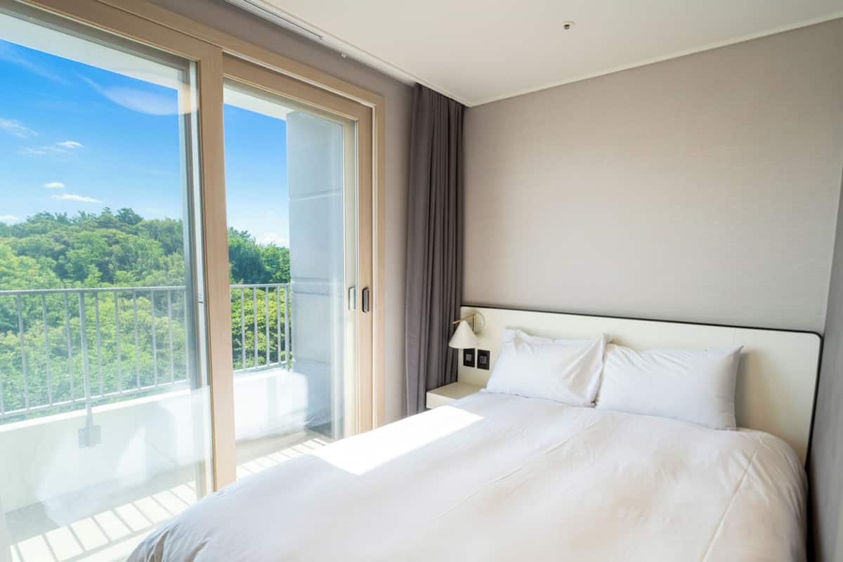 Habitación de hotel con una cama grande, una mesita de noche, una lámpara y un ventanal corredero que da a un balcón con vistas a la vegetación.