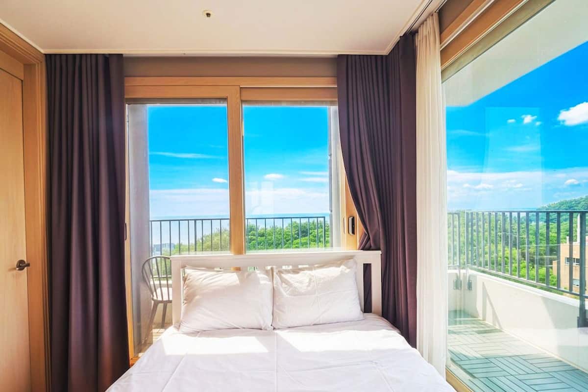 Un dormitorio con cama, una silla, un balcón con barandilla y vista a zonas verdes.