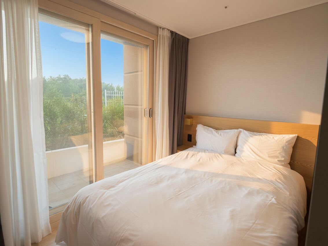 A hotel room with a bed, a view through a sliding door to an outdoor area, and a neutral-colored wall.