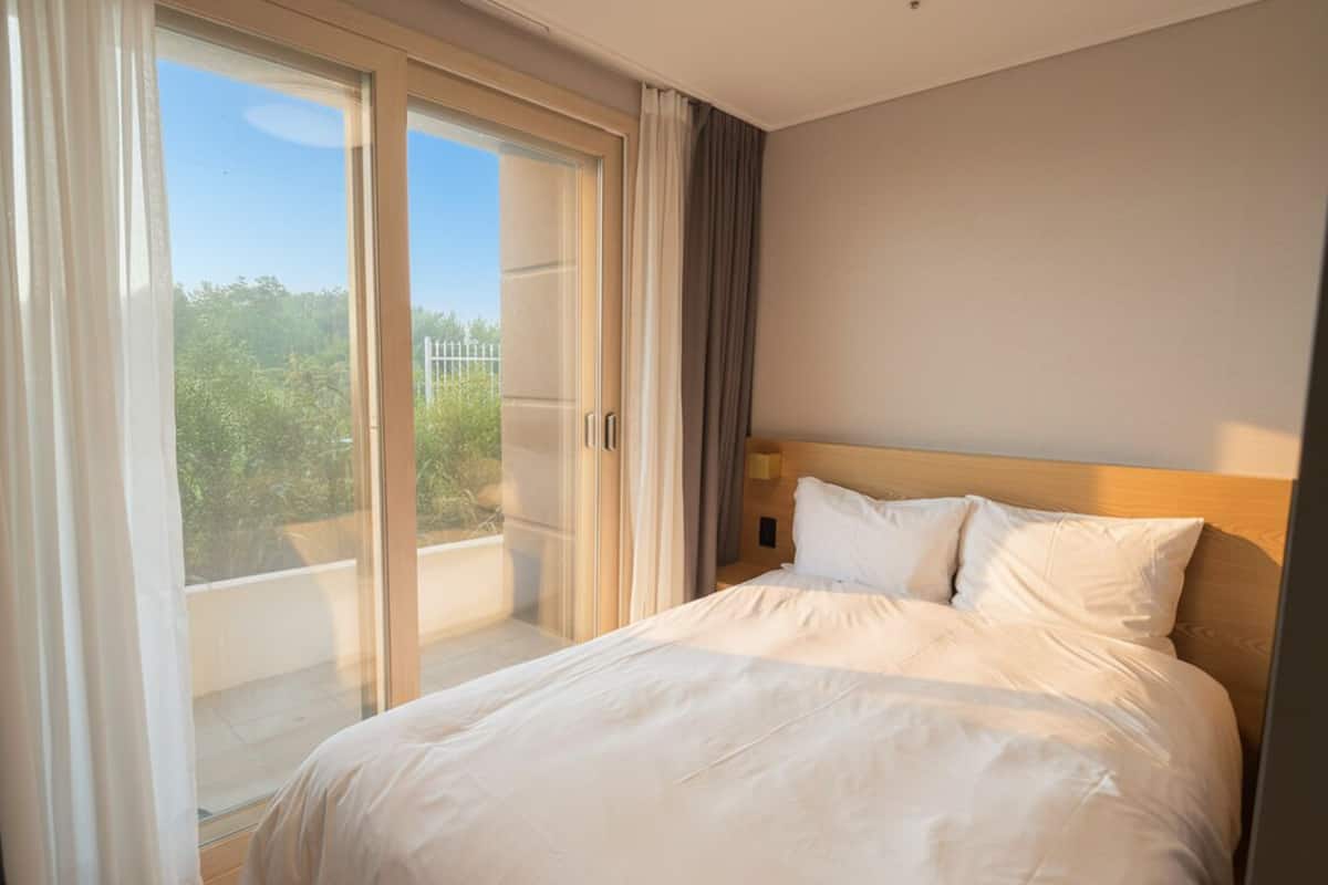 Habitación de hotel con cama, vista a un espacio exterior por una puerta corredera y pared de color neutro.