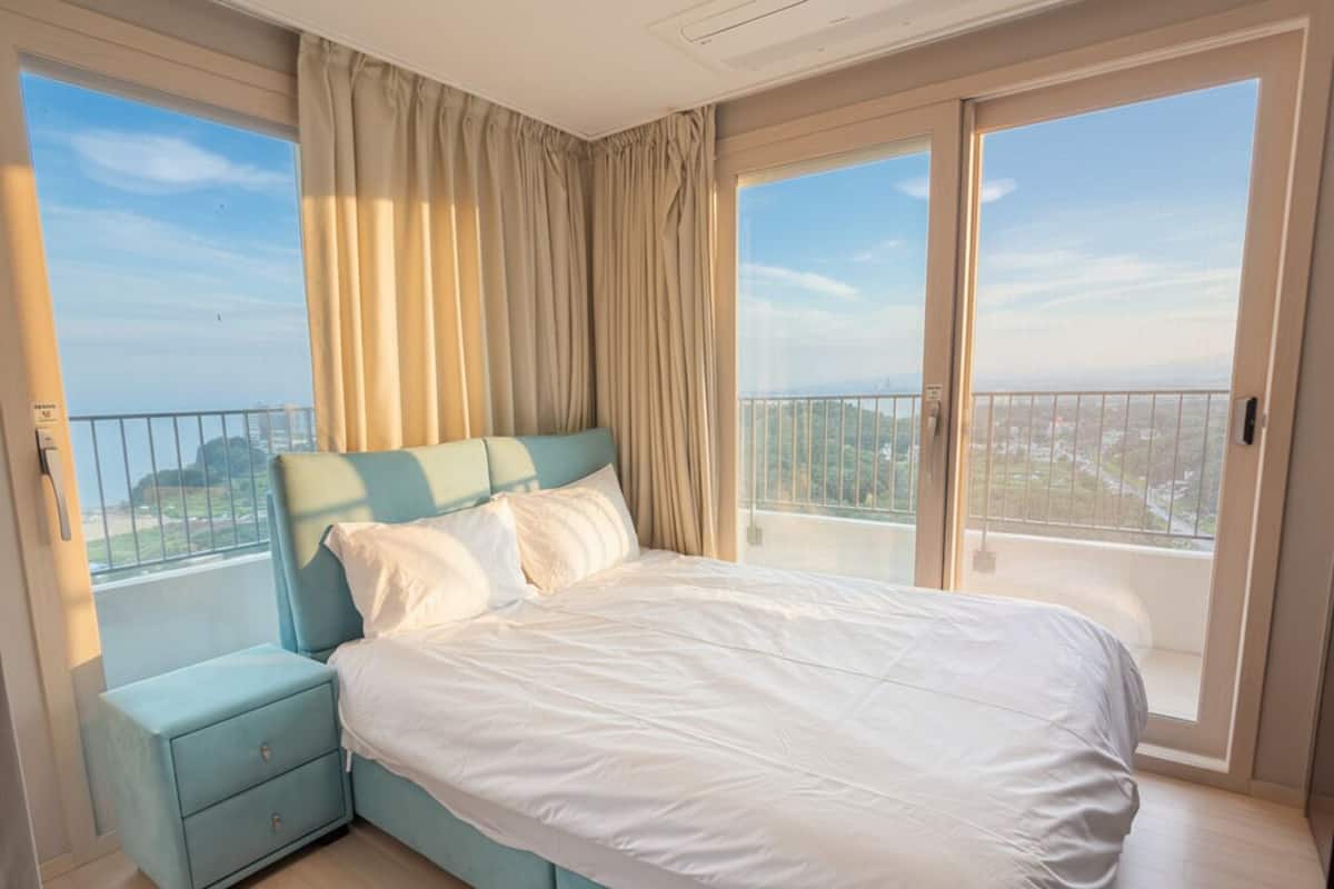 Un dormitorio con una cama grande, una mesita de noche y vistas a un paisaje urbano desde un balcón.