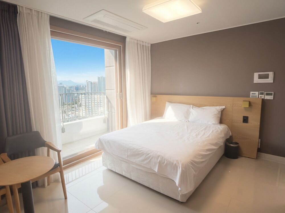 A hotel room with a large bed, a chair, a small table, a window with curtains, and a view of a cityscape.