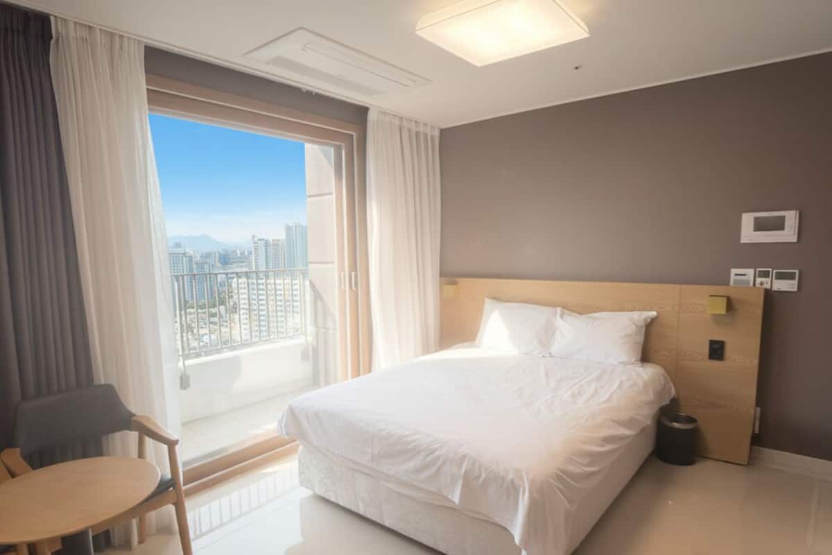 Habitación de hotel con una cama grande, una silla, una mesita, una ventana con cortinas y vistas a la ciudad.