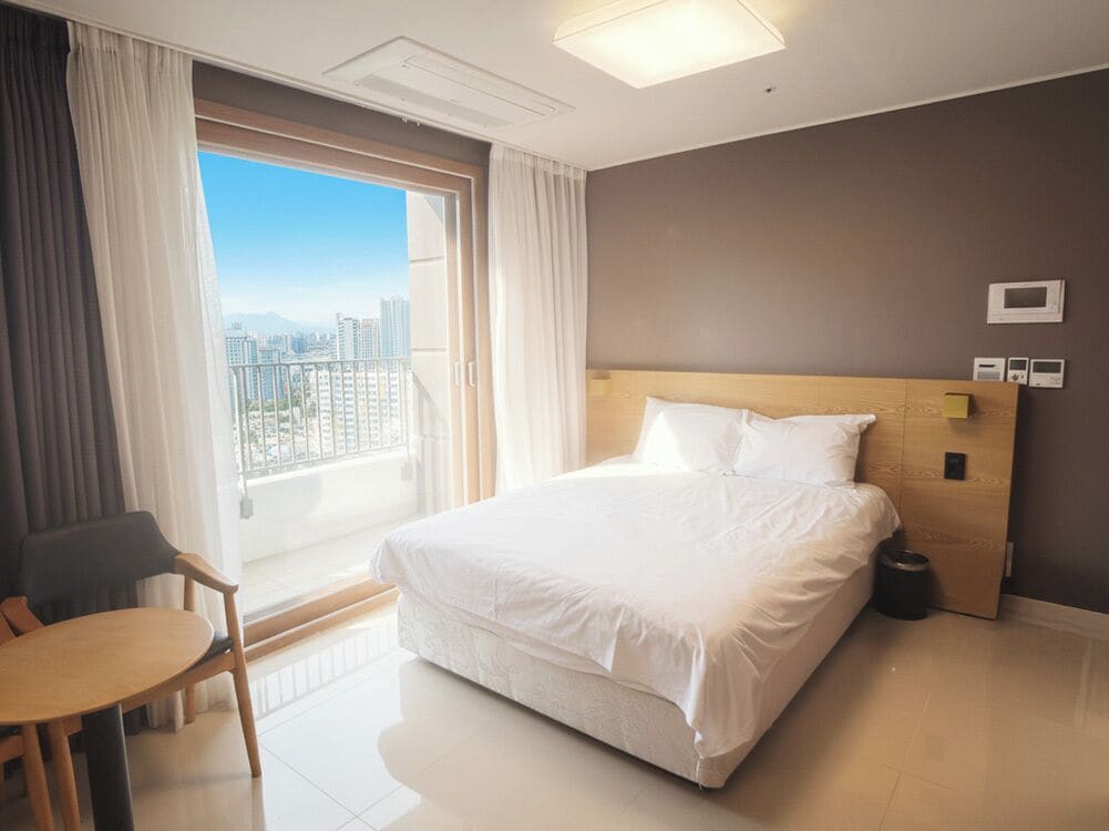 A hotel room with a large bed, a small table, a chair, a view of the city, and a window with curtains.