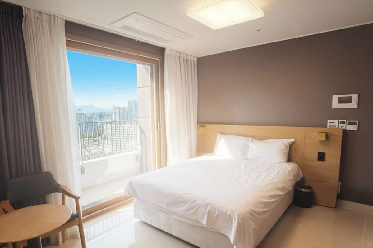 A hotel room with a large bed, a small table, a chair, a view of the city, and a window with curtains.