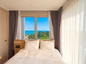 A hotel room with a bed, curtains, a window with a view of the sea, and a door.