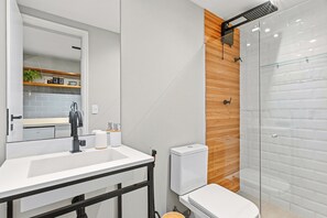 Uma casa de banho moderna com lavatório branco, espelho e sanita, chuveiro com porta de vidro e parede de destaque em madeira.
