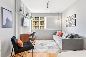 Uma sala de estar moderna com um sofá, uma poltrona, uma escrivaninha e uma janela com vista para um edifício.