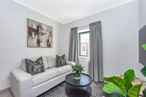 En stue med en sofa, et rundt sofabord, et billede på væggen og et vindue med gardiner.