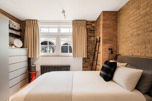 Ein Schlafzimmer mit Backsteinwand, einem Bett mit Kissen, einem Heizkörper, einer Uhr und einem Fenster mit Vorhängen.