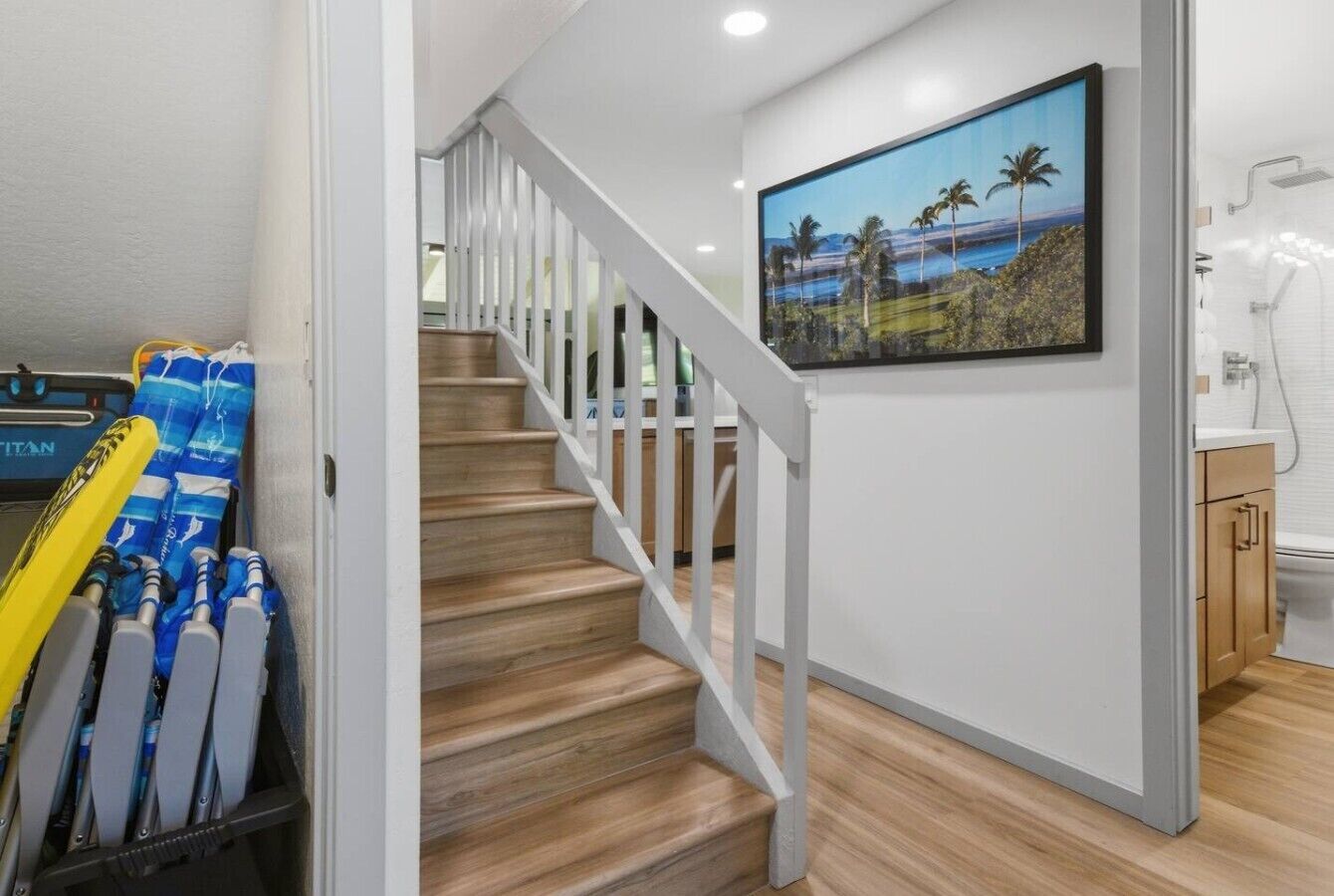 Una escalera con barandas blancas que lleva a una habitación con piso de madera, un cuadro de una playa y un baño con ducha.