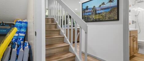 Una escalera con barandas blancas que lleva a una habitación con piso de madera, un cuadro de una playa y un baño con ducha.