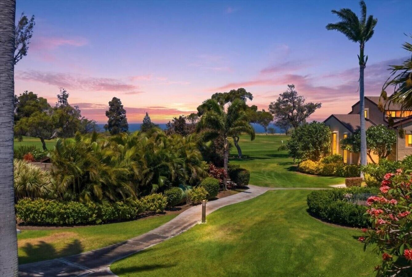 A well-maintained garden with a curved pathway leading to a building, surrounded by lush greenery and palm trees.