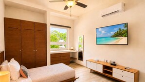 Un dormitorio con cama, armario de madera, tocador con espejo, televisor y ventana con vista a follaje.