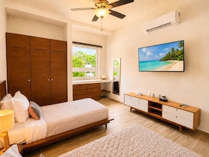 A bedroom with a bed, a wooden wardrobe, a vanity with a mirror, a TV, and a window with a view of greenery. - Boutique Tropical Escape | Pool + Beach (Puerto Aventuras)