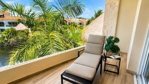 Un balcón con una silla de descanso, una mesita con una planta y vistas a una piscina y vegetación tropical.