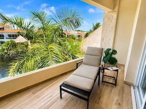 A balcony with a lounge chair, a small table with a plant, and a view of a pool and tropical vegetation. - Boutique Tropical Escape | Pool + Beach (Puerto Aventuras)