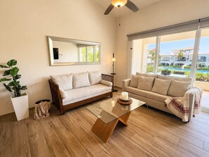 A living room with two sofas, a glass coffee table, a ceiling fan, a large window with a view of a pool and buildings, and a potted plant. - Boutique Tropical Escape | Pool + Beach (Puerto Aventuras)