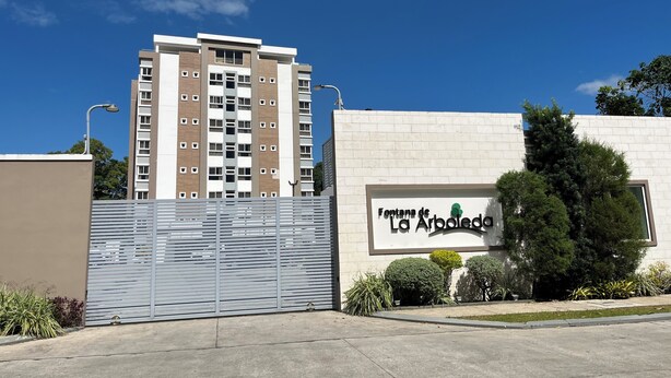 Une entrée fermée par un portail mène à un immeuble résidentiel, et une plaque indique : « Fontes de La Arboleda ».