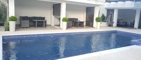 Una piscina con borde de azulejos, rodeada de zonas de estar al aire libre con mesas y sillas, y plantas en macetas.