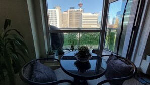 Quarto com vista para a paisagem urbana através de uma grande janela, uma mesa redonda com cadeiras e plantas em vasos.