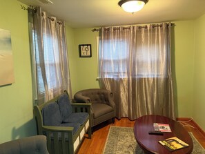 Ein Wohnzimmer mit Sofa, Sessel und Couchtisch. Vor den Fenstern hängen Vorhänge, an der Wand hängt ein gerahmtes Bild und auf dem Boden liegt ein Teppich.