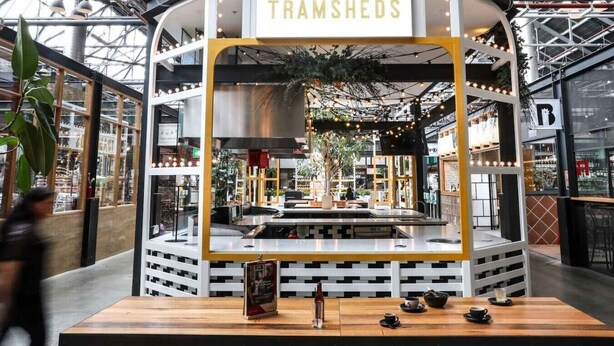Un restaurant appelé TRAMSHEDS, doté d’un intérieur moderne, d’un espace de cuisine central et offrant une vue sur le centre commercial environnant.