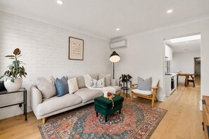 A modern living room with a white brick wall, a sofa, a green ottoman, a patterned rug, and a dining area with a table and chairs. - Charming Sunlit Terrace in Vibrant Newtown (Newtown)