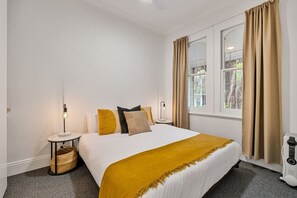 A hotel room with a bed, a bedside table with a lamp, a window with curtains, and a radiator. - Charming Sunlit Terrace in Vibrant Newtown (Newtown)