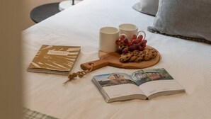 A neatly made bed with pillows, a bedside lamp, two mugs, a book, and a plate with cookies and grapes.