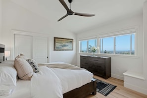 A bedroom with a bed, a dresser, a ceiling fan, a window with a view, and a painting on the wall. - Ocean & City View Retreat on the Riviera (Santa Barbara)