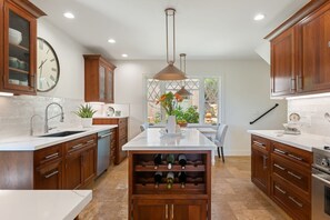 A modern kitchen with wooden cabinets, a central island with wine bottles, a clock, and a pendant light. - Ocean & City View Retreat on the Riviera (Santa Barbara)