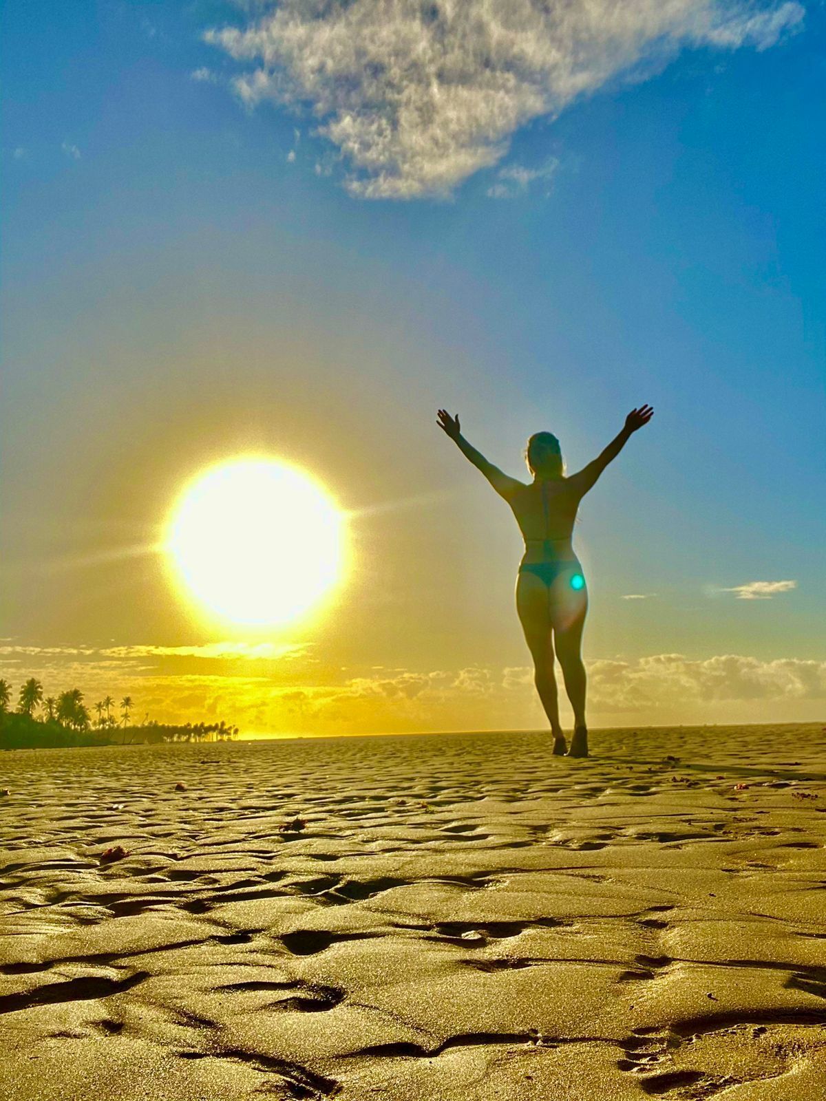 Una persona de pie en la playa, con los brazos levantados, mirando al sol.
