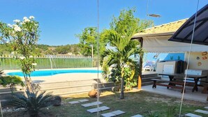 A poolside area with a clear view of the pool, a covered seating area, and a patio with stone tiles and potted plants. - In Paradiso  (Aracruz)