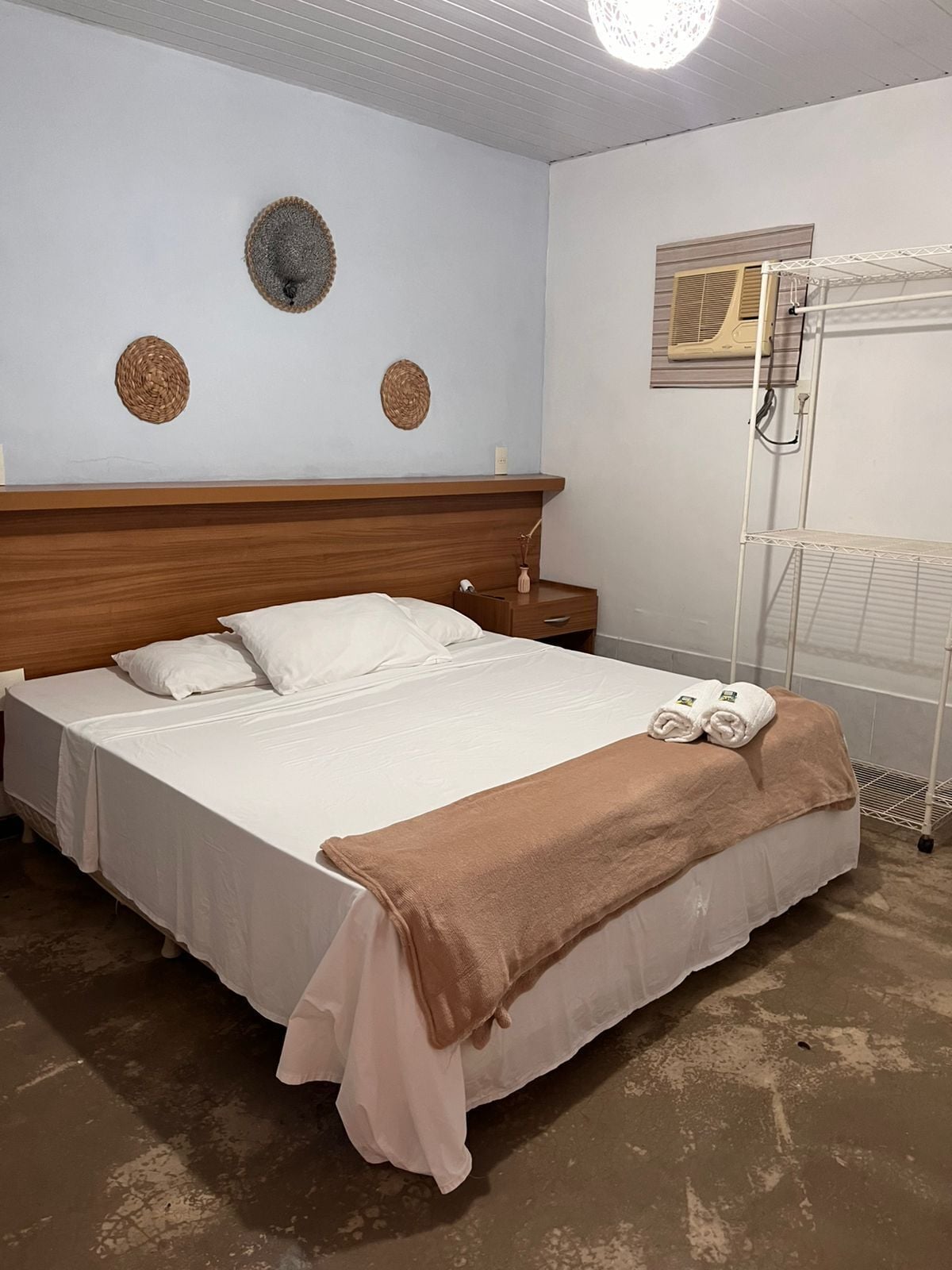 Un dormitorio con cabecera de madera, cama con sábanas blancas, una manta marrón y una mesita de noche con lámpara.